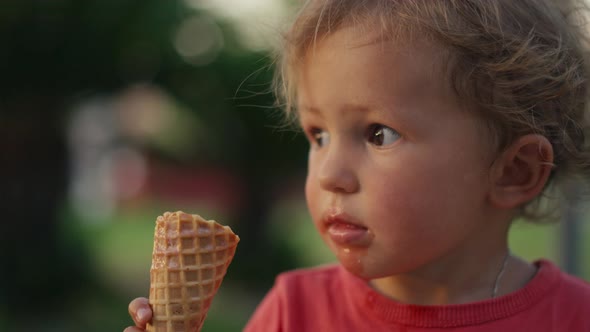 Fairhaired Boy Eats White Cold Cream Ice Cream in Waffle Cone Cup While Sitting Outside in Sunny