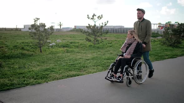 Man Is Rolling Wheelchair with Young Blonde Woman Walking Together in Park