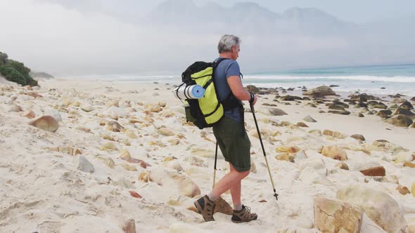 Senior hiker man with backpack and hiking poles walking while hiking on the beach.