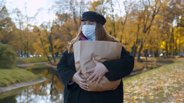 Overweight Young Woman in Safety Mask Carrying Paper Bag with Groceries Walking in Autumn Park