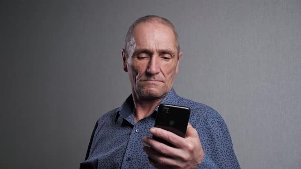 Senior Man Reads News in Mobile Phone on Grey Background