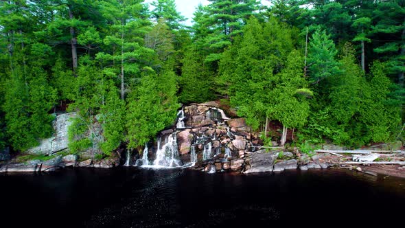 Water cascading down rocky waterfall between lush green forest into a dark colored lake view from ab