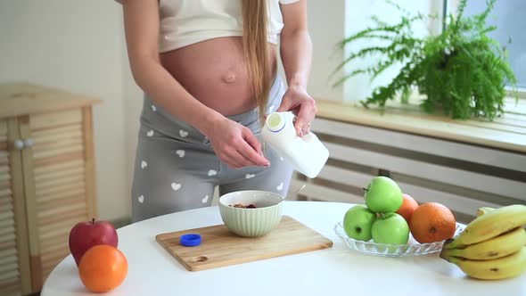 Pregnant Woman Preparing Muesli with Milk and Standing at Table in Home Kitchen Spbd