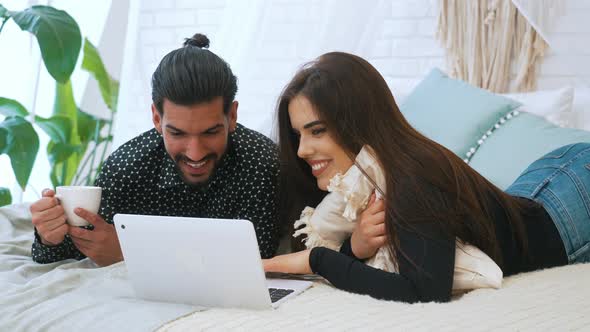 Young Happy Smiley Millennial Couple on Bed Watching Favorite Show on Silver Laptop