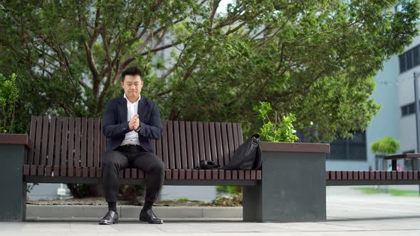 Asian businessman in formal suit worried and anxious sitting on a bench in a city park outside.