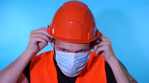 Male Construction Worker in Overalls Putting on Medical Mask on Face on Blue Background