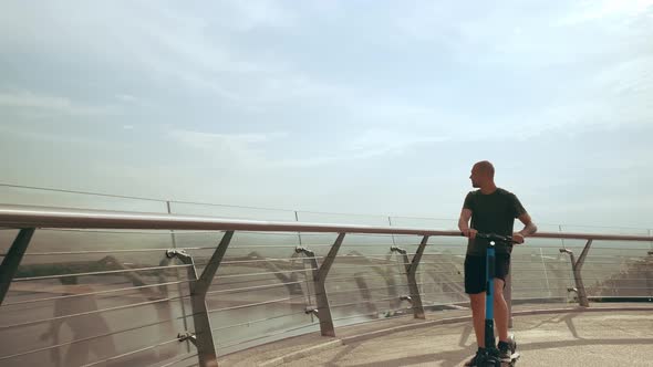 Modern Young Man Rides an Electric Scooter Across the Pedestrian Bridge Overlooking the City