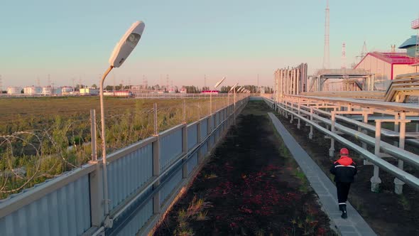 A Drone Flies Behind an Employee in an Industrial Area Behind a Fence with Barbed Wire Pipes with