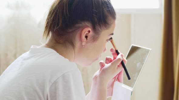 a Young Teenage Girl Makes Herself Makeup with a Small Mirror in the Bedroom
