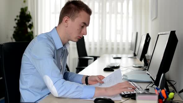 Young Handsome Man Works on Desktop Computer and Reads Document in the Office