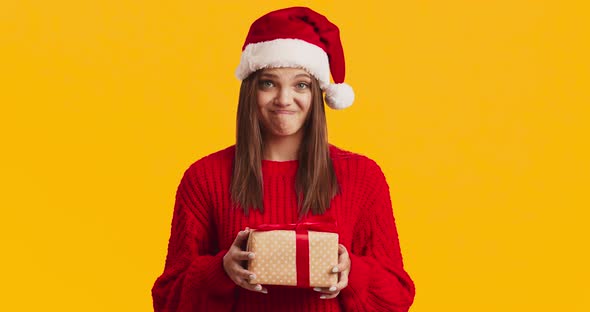 Young Displeased Woman in Santa Hat Holding Present Box and Frowning, Orange Studio Background