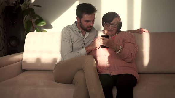 Grandson Teaching Grandma Using Mobile Phone Camera. Grandson And Grandmother Sitting On Sofa.