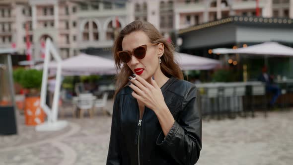 a Woman with Bright Lipstick and Sunglasses Walks with a Cigarette in Hand Through the City Square