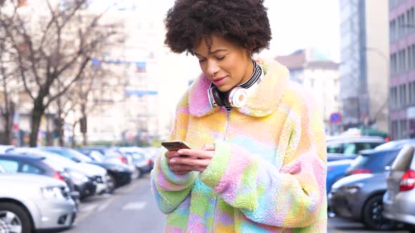 Young beautiful curly hair black woman outdoor in the city smiling using smartphone