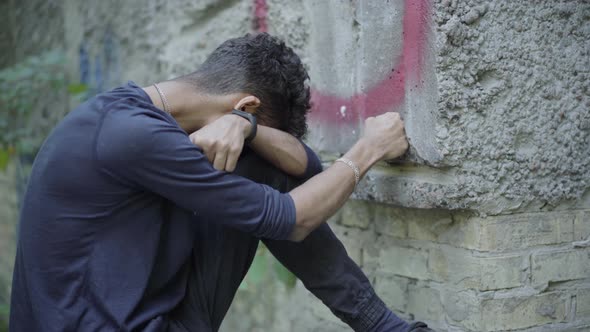 Desperate Lonely Young Man Punching Wall with Fist and Crying. Side View of Frustrated Stressed