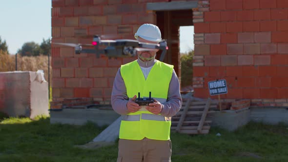 Drone Operator Holding Remote Controller Controls an Aircraft in Front of Construction House Site