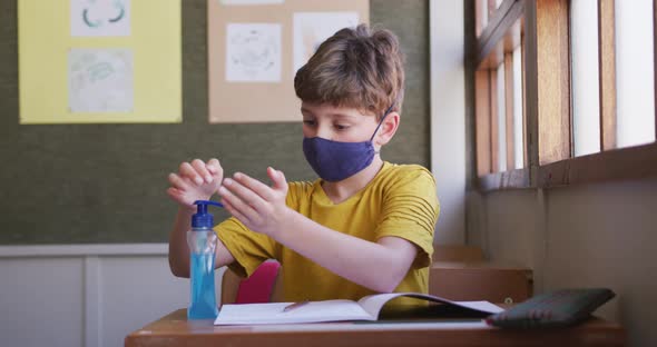 Boy wearing face mask sanitizing his hands while sitting on his desk at school