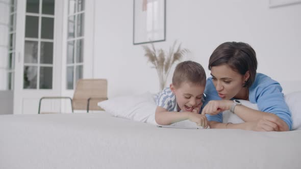 Happy Young Mother Showing Cartoon on Digital Tablet to Her Toddler Son Before Going to Sleep