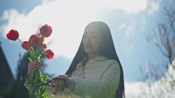 Young Smiling Asian Woman Cutting Flower Stems with Scissors Standing in Sunlight with Blued Cloudy