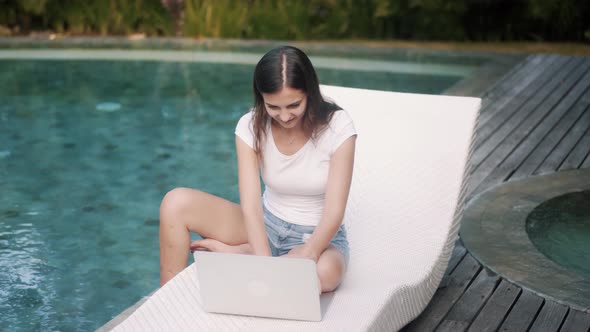 Joyful Girl Works on Laptop Sitting on Couch Near Pool