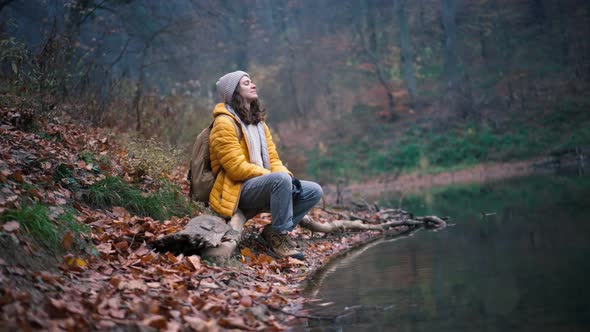 A Young Woman Sits on the Shore of a Forest Pond and Enjoys the Fresh Air