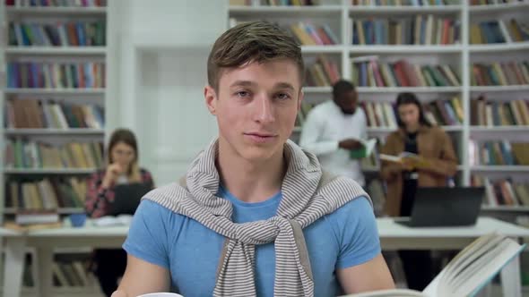 Smiling Modern Young Man which Reading Book in the Library and Looking at Camera