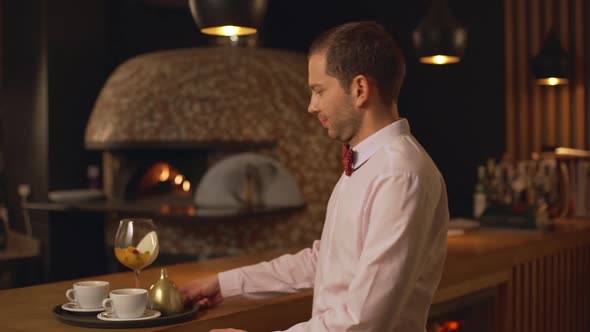 Middleaged Caucasian Male Waiter in White Shirt and Bow Tietaking Tray with Cups and Glass Smiling