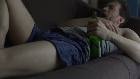 Guy in Boxers and Tank Top Dozing on Sofa, Beer Bottle in His Hand, Alcoholism