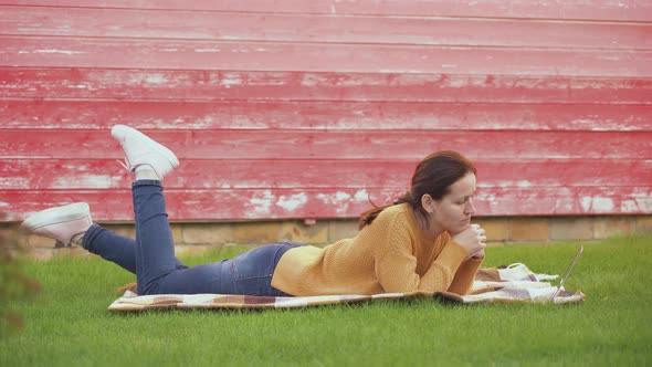 A girl with a laptop is lying on a blanket on the lawn and communicating via video link