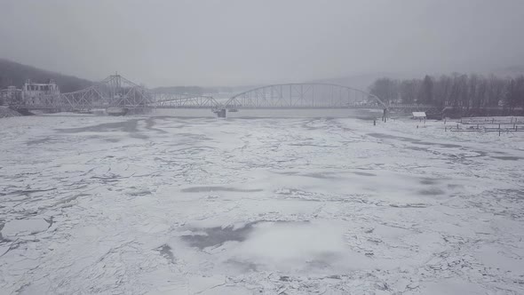 Drone flying towards swing bridge above a snowy river with big ice chunks from a winter storm.