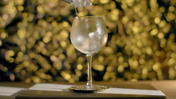 Ice Cubes are Being Dropped to the Cocktail Glass in Slow Motion on the Golden Sparkle Background