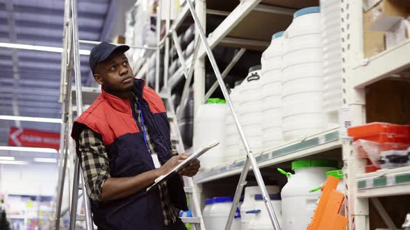 Male Supervisor Examining Goods on Shelves with Tablet Using Ladder