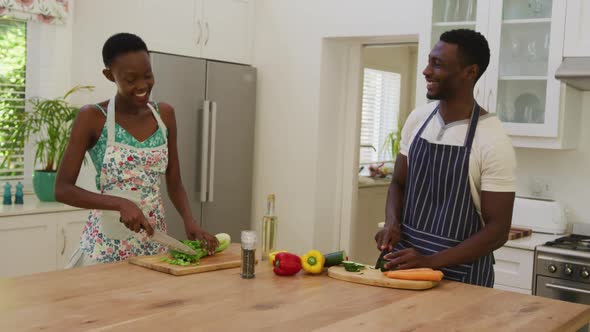 Smiling african american couple wearing aprons talking and preparing food in kitchen