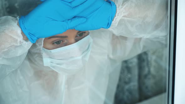 Sick isolated pensive girl in protective suit, face mask, gloves stares longingly out through window