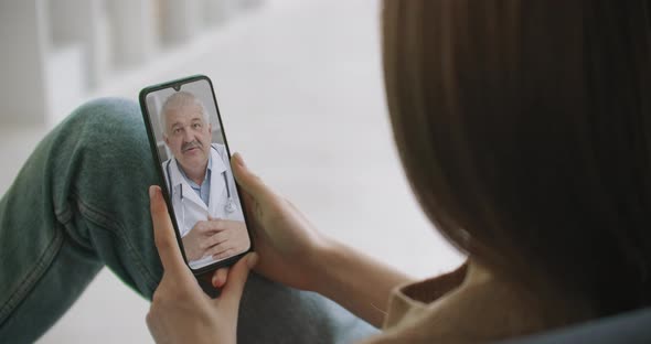 Female Medical Assistant Wears White Coat, Headset Video Calling Distant Patient on Smartphone