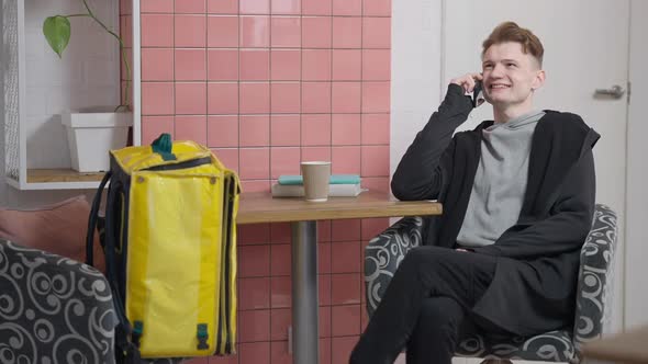 Laughing Relaxed Young Caucasian Man Talking on Smartphone Sitting in Cafe with Yellow Food Delivery