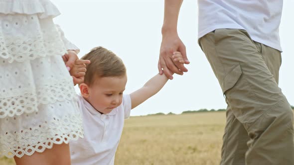 Toddler Boy Walks Holding Hands of Mother Father in Field