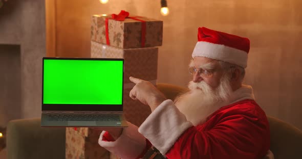 Santa Holds a Laptop with a Green Screen While Sitting in the Christmas Decorations