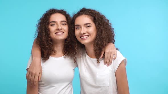 Two Charming Caucasian Curly Female Twins in White Tshirts Embracing Each Other on Shoulders Smiling