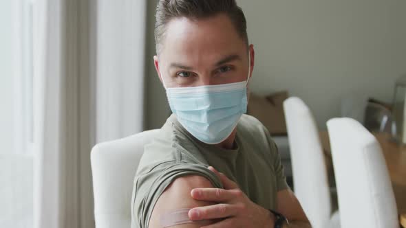 Portrait of caucasian man wearing face mask showing his vaccinated shoulder at home