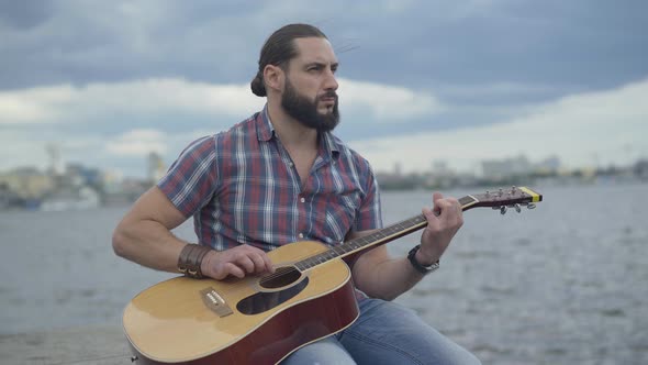 Brunette Caucasian Bearded Man Playing Guitar with Blurred City at the Background. Confident Young