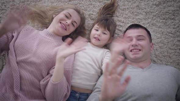 Top View Close-up of Smiling Caucasian Family Lying on Soft Carpet and Waving at Camera 
