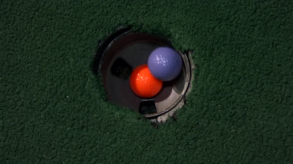 A close up birds eye view of an orange ball in a hole and then a purple and yellow golf ball fall in