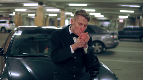 Young Man in a Black Suit with a Bowtie Smoking a Cigarette Sitting at the Bonnet of the Black Car