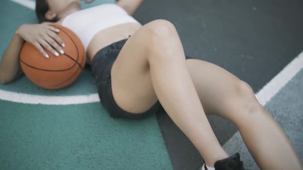 Camera Moves Along Fit Young Caucasian Woman Lying on Basketball Sports Ground with Orange Ball and