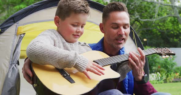 Happy caucasian father with son sitting in tent and playing guitar in garden