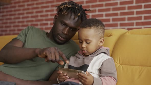 Portrait of African American Father Sitting with Son on Sofa  Kid Using Smartphone