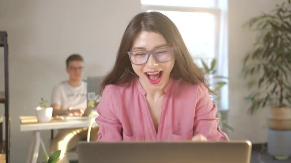 Beautiful Asian Businesswoman in Glasses Sitting at Her Office Desk in Front of Laptop Raising Arms