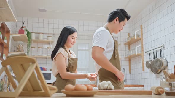 Attractive wife tying apron laces to husband with love, spend time together in kitchen at home.