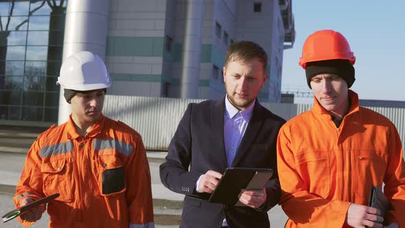 Engineer in a Suit and Two Workers in Orange Uniform and Helmets are Walking Through Building